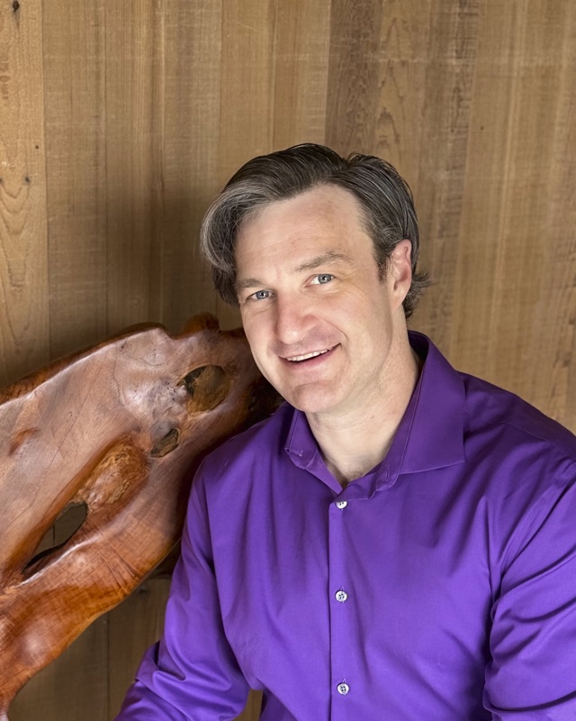Ben Stull, smiling, in a purple shirt beside a carved wood sculpture